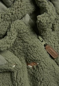 Olivgrön fleecejacka med texturerad yta, dragkedjestängning och lädermärke. Har tryckknappar och en mjuk innerfoder.