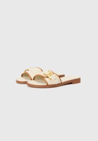 Beige slip-on sandals with two adjustable straps and gold hardware accents. Features a flat, brown sole and soft, textured footbed.