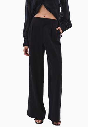 Vrouw met een hoge taille, zwarte broek met wijde pijpen en een transparante zwarte blouse met lange mouwen, staand met één hand in de zak.