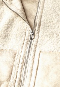 Veste zippée beige avec une texture en fausse fourrure à l'avant et un tissu lisse à l'arrière, dotée d'une fermeture éclair en métal argenté.