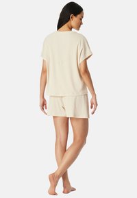 Frau, barfuß stehend, von hinten zu sehen, trägt ein hellbeiges Kurzarm-Top und passende Shorts vor einem schlichten hellen Hintergrund.