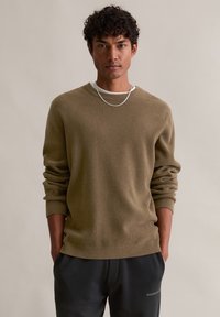 Pull en maille vert olive avec des manches longues, un col rond et une coupe décontractée, porté sur une chemise blanche, associé à un pantalon foncé. Chaîne en argent simple.