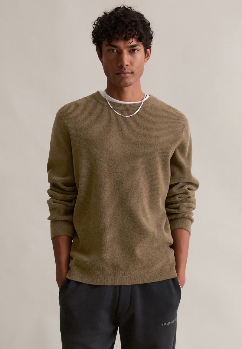 Pull en maille vert olive avec des manches longues, un col rond et une coupe décontractée, porté sur une chemise blanche, associé à un pantalon foncé. Chaîne en argent simple.