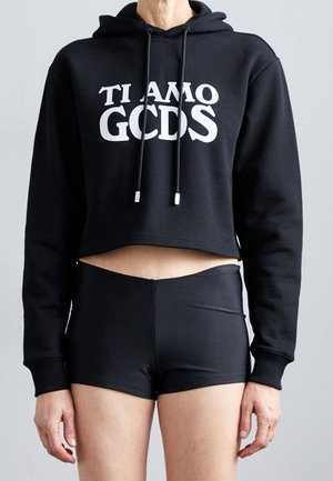 Osoba ubrana w czarną krótką bluzę z kapturem z napisem "TI AMO GCDS" w białej czcionce oraz czarne szorty z wysokim stanem, stojąca na jednolitym tle.