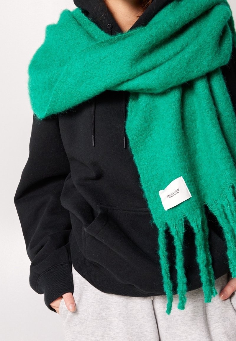Personne portant un sweat à capuche noir et un pantalon gris avec une grande écharpe en laine verte enveloppée autour du cou, arborant une étiquette de marque blanche.