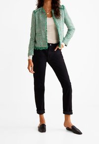 Vihreä tweed-takki, jossa hapsureunaiset yksityiskohdat, valkoinen istuva toppi, mustat housut taitettavalla helmalla ja mustat nabarit. Yksinkertainen, teksturoitu muotoilu.