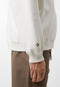 Sudadera crema con puños acanalados, que presenta un pequeño logotipo en forma de estrella marrón en la manga izquierda y un corte relajado. Textura de algodón suave.