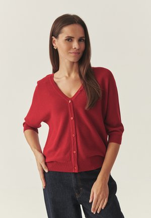 Femme aux longs cheveux bruns portant un cardigan rouge boutonné et un jean foncé, debout avec une main sur la hanche devant un fond uni.
