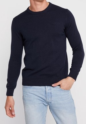 Mand iført en tætsiddende mørk marineblå langærmet sweater med lyseblå jeans, den ene hånd i lommen og den anden afslappet ved siden.