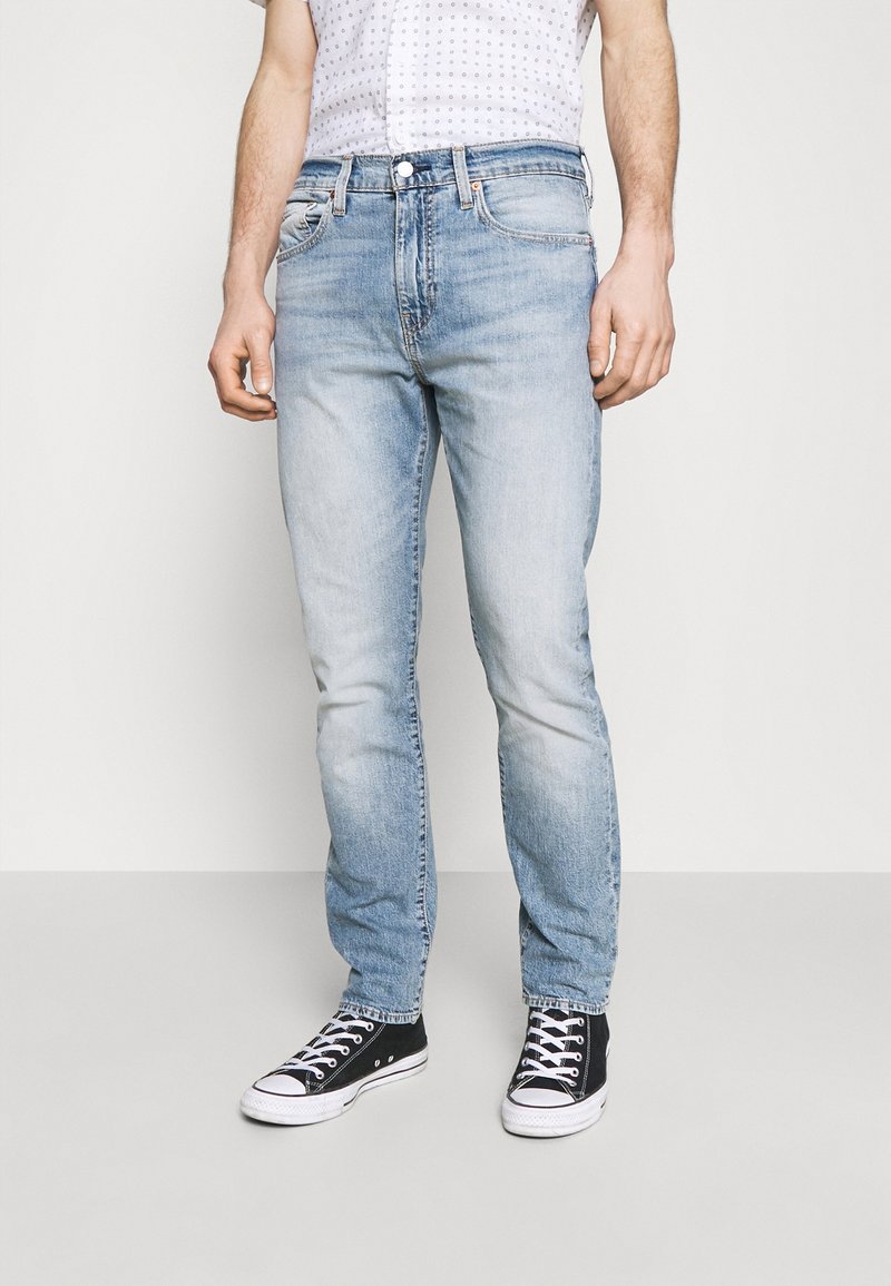 Homme portant un jean délavé bleu clair, des baskets noires et blanches, et un t-shirt blanc à motifs, debout devant un fond uni.