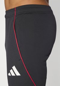 Pantalones cortos deportivos negros con un diseño ajustado, que cuentan con una franja lateral roja y un logo blanco en la parte inferior izquierda. Textura de tela suave.