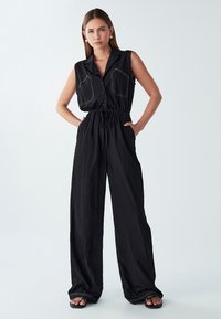 Ženska v črnem brezrokavnem kombinezonu z vrvico v pasu, širokimi hlačami, sprednjim gumbom in kontrastnimi šivi na prsnih žepih, stoji z rokami v žepih.