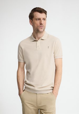 Mężczyzna o jasnej karnacji i krótkich brązowych włosach, ubrany w beżową koszulkę polo i beżowe spodnie, stoi z rękami w kieszeniach, patrząc w lewo.