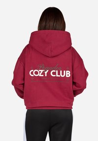Bordová mikina s voľným strihom, na zadnej strane s veľkým bielym a zeleným potlačou, vrátane nápisu "Cozy Club" a ďalších textových detailov.