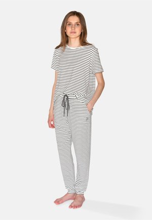 Junge Frau steht barfuß und trägt ein passendes weiß-schwarz horizontal gestreiftes kurzärmeliges Oberteil und eine Hose mit Kordelzug.