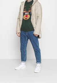 Beige jas met een kraag aan de binnenkant van bont, groene trui met een rendierontwerp, blauwe spijkerbroek en witte sneakers. Casual outfit met speelse grafische elementen.
