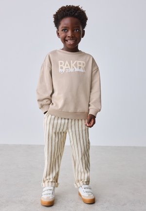 Giovane ragazzo sorridente che indossa una felpa beige "BAKER", pantaloni a righe e sneakers bianche, in piedi con le mani parzialmente nelle tasche.