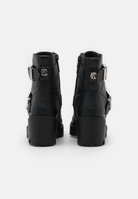 Stivaletti in pelle nera con due fibbie argento sui lati, suole in gomma spesse e parte superiore testurizzata. La vista posteriore mostra le cerniere.