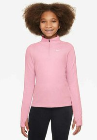 Ragazza con capelli ricci che indossa un top a maniche lunghe rosa con zip a quarto di Nike e pantaloni neri, in piedi e sorridente contro uno sfondo bianco.