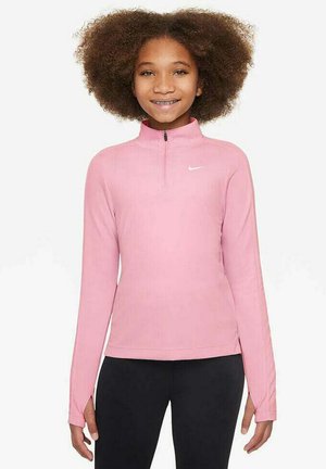 Ragazza con capelli ricci che indossa un top a maniche lunghe rosa con zip a quarto di Nike e pantaloni neri, in piedi e sorridente contro uno sfondo bianco.