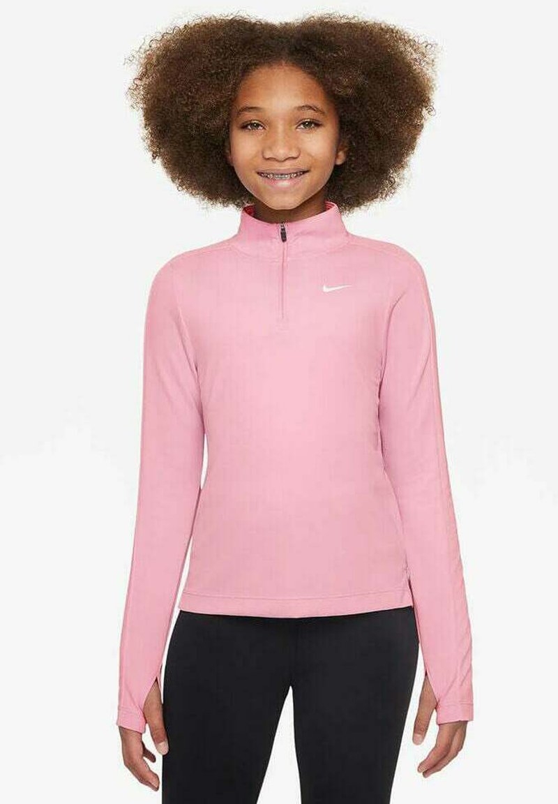 Ragazza con capelli ricci che indossa un top a maniche lunghe rosa con zip a quarto di Nike e pantaloni neri, in piedi e sorridente contro uno sfondo bianco.