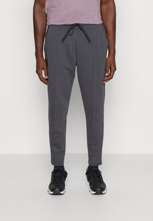 Uomo che indossa pantaloni della tuta grigio scuro a gamba stretta con coulisse in vita e sneakers nere, in piedi contro uno sfondo chiaro.