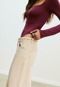 Bordeaux lange mouwen bodysuit gecombineerd met lichtbeige high-waisted jeans met decoratieve kraaldetails langs de tailleband.
