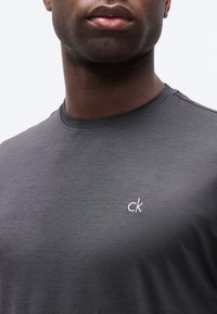 Κοντινό πλάνο ενός άνδρα που φοράει σκούρο γκρι μπλουζάκι με στρογγυλή λαιμόκοψη και μικρό λευκό λογότυπο "ck" στο αριστερό στήθος.