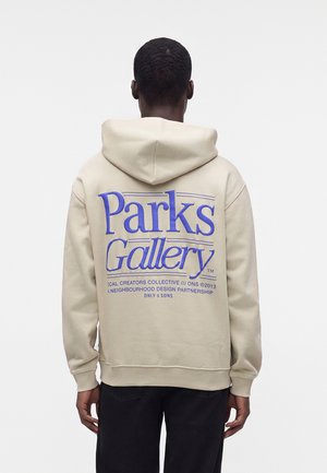 Osoba w beżowej bluzie z kapturem z dużym niebieskim tekstem "Parks Gallery" na plecach, stojąca na jednolitym jasnym tle.