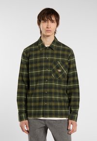Een jonge man staat in een groen ruitjesoverhemd met een borstzak, gecombineerd met grijze broek. Hij staat tegenover de camera tegen een lichte achtergrond.