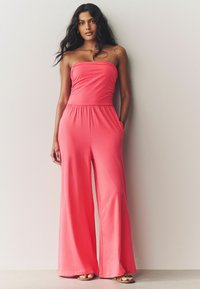 Mono sin tirantes color coral con pierna ancha, cinturilla elástica y bolsillos. Hecho de tela suave, tiene un ajuste casual.