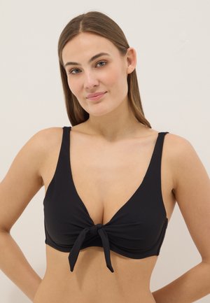 Joven mujer con cabello castaño liso lleva un top de bikini negro con nudo delantero, de pie frente a un fondo beige liso.