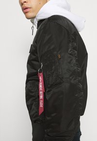 Giubbotto bomber nero in nylon con orlo a costine, tasca con zip sulla manica e un'etichetta rossa "RIMUOVERE PRIMA DEL VOLO". Felpa bianca sotto.