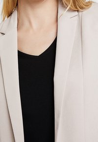 Blazer beige clair en tissu texturé, avec des revers crantés. Porté sur un haut noir à col en V, mettant en valeur une tenue superposée.