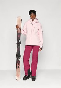 Sieviete, tērpusies rozā slēpošanas jakā, purpura slēpošanas biksēs, melnās slēpošanas zābakos un ar brillēm, tur vienu vertikāli nostiprinātu slēpi pret vienkāršu fonu.