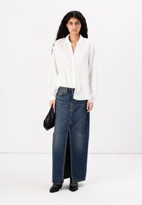 Blusa blanca con textura y patrón de diamantes; combinada con una falda larga de denim oscuro con una abertura frontal; bolso negro; zapatos negros.