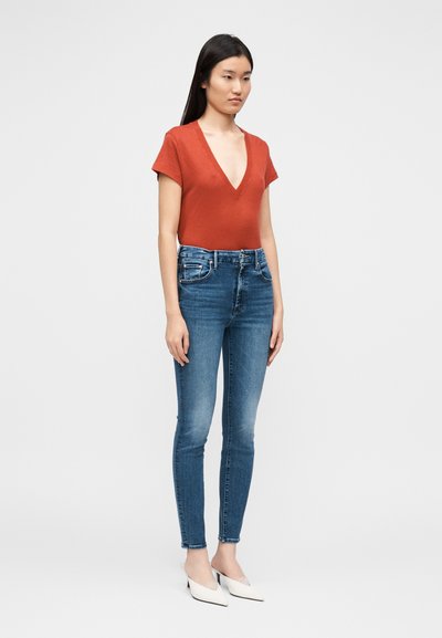 Punane lühikeste varrukatega ülemine osa sügava V-kaelusega, kombineeritud siniste kitsaste teksade ja valgete terava ninaga kontsakingadega. Tekstuur tundub sile; teksadel on veidi pleekinud koht.