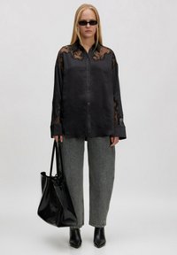 Schwarze Satinschirt mit Spitze an den Ärmeln, kombiniert mit hoch geschnittenen grauen Jeans und schwarzen Stiefeln mit Absatz. Hält eine schwarze Tote-Tasche.