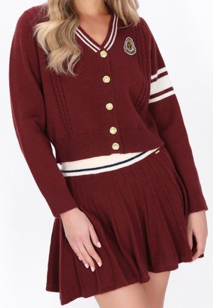 Femme portant un cardigan en maille bordeaux avec des boutons dorés et une jupe plissée assortie, avec des détails à rayures blanches et bleu marine.