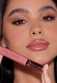 Płynny błyszczyk do ust w różowym opakowaniu, z błyszczącą złotą nakrętką. Modelka trzyma produkt, prezentując gładkie, połyskujące wykończenie na ustach.