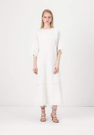White cotton maxi dress with short flared sleeves, textured vertical stripes, and lace accents. Worn with brown strappy heels.
