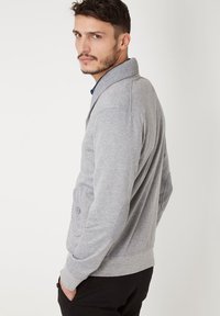 Jersey gris con cuello de chal, acentos acanalados y bolsillos con botón. La tela parece suave con un diseño ajustado, usado sobre una camisa azul.
