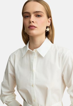 Mujer con cabello rubio liso lleva una camisa blanca abotonada y grandes pendientes dorados con forma de estrella, mirando ligeramente hacia un lado.
