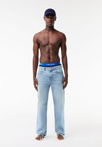 Jeans bleu clair à coupe décontractée, avec une ceinture Lacoste bleue et une casquette navy unie. Le modèle est torse nu et pieds nus.