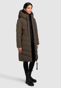 Abrigo acolchado color verde oliva con capucha, presenta un diseño acolchado, bolsillos laterales con cremallera y cordones ajustables. Lleva puesto sobre pantalones negros y botas.