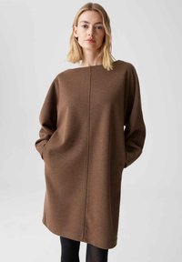 Bruine oversized jurk met een ronde halslijn, lange mouwen en een centrale naaddetail, gemaakt van een zachte, gestructureerde stof.