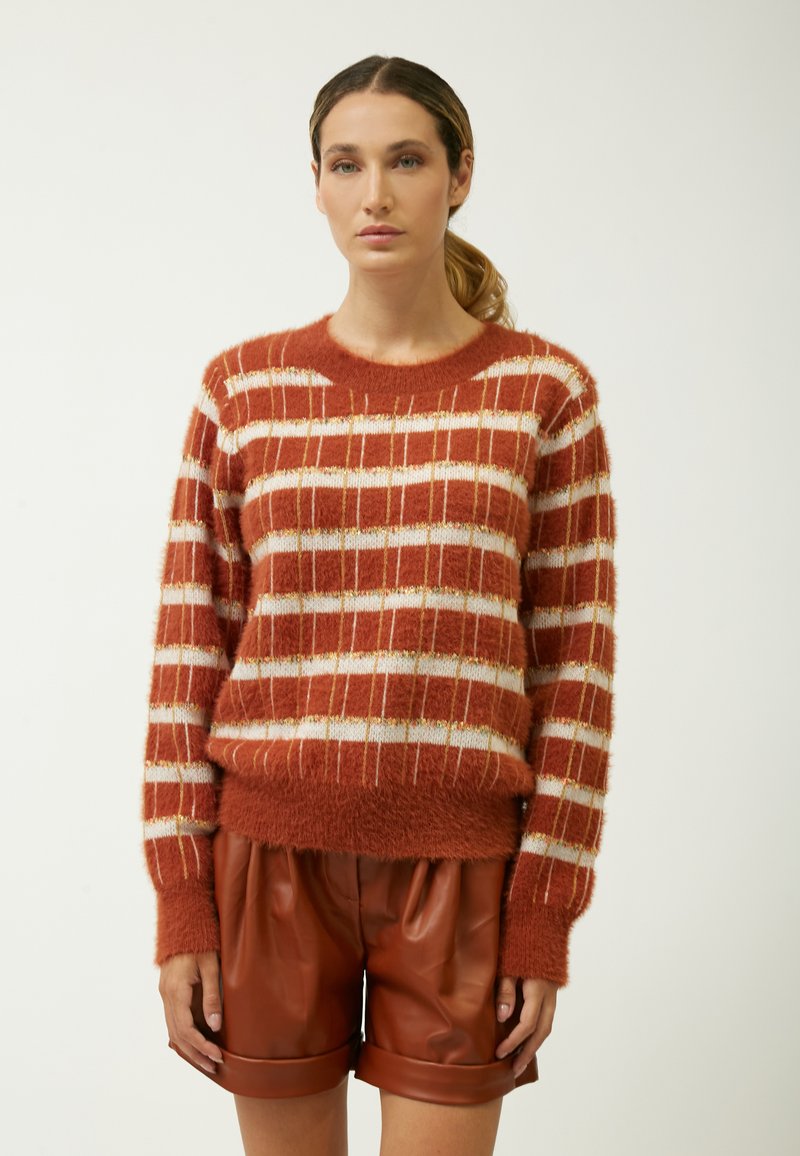 Strickpullover in Rostrot mit weißen und metallisch goldenen Streifen, weichem Material und rundem Ausschnitt, kombiniert mit braunen Kunstleder-Shorts.