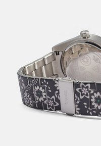 Silberner Edelstahl-Uhr mit einem gemusterten schwarzen Armband, das weiße und bunte Blumen zeigt und eine Kombination aus Materialien und Texturen präsentiert.