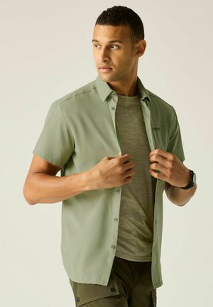 Uomo che indossa una camicia verde chiaro a maniche corte abbottonata sopra una t-shirt grigia, che sistema la camicia guardando leggermente di lato.