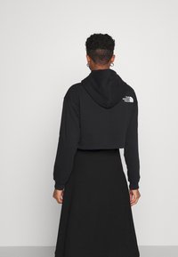 Černá zkrácená mikina s kapucí z měkkého materiálu, s kapucí a malým bílým logem na zádech. Kombinováno s dlouhou černou sukní.